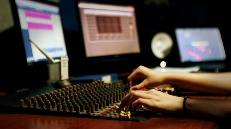 A recording engineer sitting at a console while tracking a vocalist in a vocal booth.